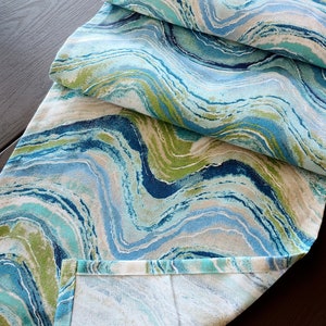 May include: A decorative table runner with a swirling pattern in shades of blue, green, and white. The fabric has a marbled effect, creating a flowing, organic design. The runner is folded over, showing the hemmed edge.