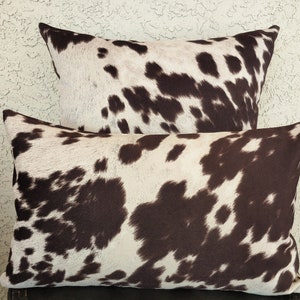 May include: Two decorative throw pillows with a brown and white cowhide pattern. The pillows are stacked on top of each other.