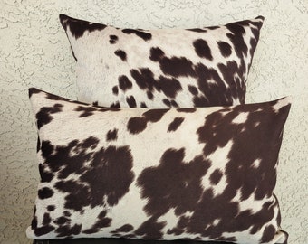 May include: Two decorative throw pillows with a brown and white cowhide pattern. The pillows are stacked on top of each other.