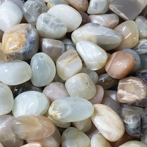 May include: A collection of smooth, polished moonstone tumbled stones in various shades of white, gray, and brown. The stones have a natural, iridescent sheen.