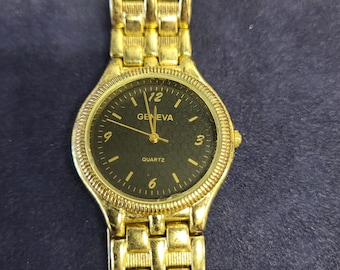 May include: Gold-toned Geneva quartz watch with a black dial. The watch features gold hour markers and hands, with the word "Geneva" printed above the center. The watch band is also gold-toned and has a linked design.