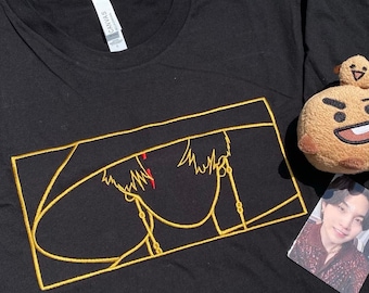 May include: Black t-shirt featuring a gold embroidered design of a face with red accents. The design is framed in a gold rectangle. A brown plush toy and a photo card are also visible.