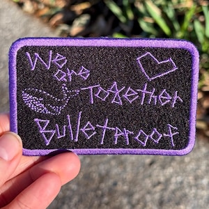 Könnte beinhalten: Ein rechteckiger schwarzer Aufnäher mit lila Nähten und Rand. Der Aufnäher zeigt den gestickten Text "We are Together Bulletproof" in Lila, zusammen mit einem Herz und einem Wal-Design.