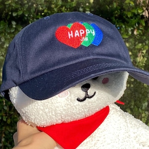 Puede incluir: Una gorra de béisbol azul marino con un diseño de corazón rojo, verde y azul que dice "Happy Jin". La gorra se lleva sobre un juguete de peluche blanco con una bandana roja.