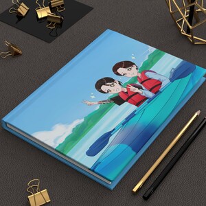 May include: A light blue journal with a vibrant illustration of two people kayaking on the cover. The artwork features a blue kayak, a scenic background, and the figures wearing life vests. Gold and black pencils and paper clips are also visible.