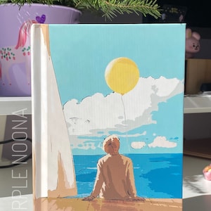 May include: A light blue and white notebook with a painting of a person sitting on a wooden deck overlooking a blue ocean. A yellow balloon floats in the sky above the person. The notebook has the words "PURPLE NOONA" printed on the side.
