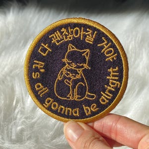 May include: A round, embroidered patch with a black background and gold trim. The patch features a cat illustration and text in Korean and English that reads "It's all gonna be alright."