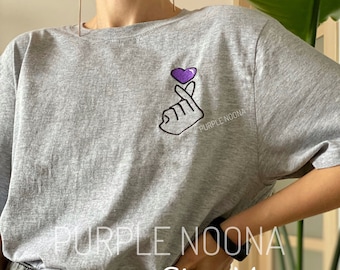Pode incluir: Camiseta cinza com um coração roxo e um gesto de mão bordados na frente. O texto "PURPLE NOONA" está bordado abaixo do coração. A camiseta é tamanho M.