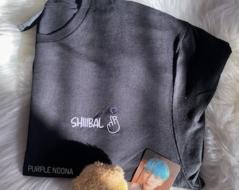 May include: Black t-shirt with white embroidery that says "SHIIIBAL" and a pink heart with a finger heart gesture. The shirt is on a white fluffy surface with a small brown teddy bear and a photo card of a person with blue hair.