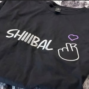以下が含まれることがあります： 白い文字で「SHIIIBAL」と書かれた黒いTシャツ。白い手のジェスチャーと、その上に紫色のハートが描かれている。