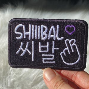 Peut inclure: Écusson rectangulaire noir avec le texte brodé blanc "SHIIIBAL" en anglais et en caractères coréens. Un cœur violet et un geste de la main sont également brodés sur l'écusson.