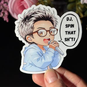 May include: A sticker featuring an anime-style character with glasses and a microphone. The character is wearing a blue hoodie and has a speech bubble that says "DJ, SPIN THAT SH*T!". The sticker has a white border.