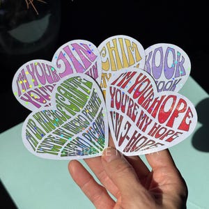 Puede incluir: Una colección de pegatinas en forma de corazón con detalles holográficos. Cada pegatina presenta un color y texto diferentes, incluyendo frases como "I'm Your Hope" y "You're My Hope". Las pegatinas se sostienen en una mano.