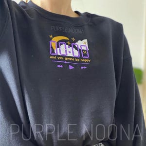 Puede incluir: Sudadera negra con un diseño gráfico púrpura. El diseño incluye el texto "and you gonna be happy" y el nombre de la marca "PURPLE NOONA". El gráfico es una interfaz de reproductor de música estilizada.