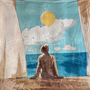 May include: A beige and brown throw blanket with a printed image of a person sitting on a wooden deck overlooking a blue ocean with a yellow sun and white clouds in the sky.