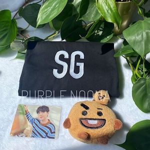May include: A black t-shirt with the letters "SG" embroidered in white on the front. The t-shirt is lying on a white surface next to a brown plush toy with a face and a photo card of a person.
