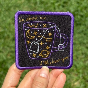 Może przedstawiać: Haftowana naszywka z czarnym tłem i fioletową obwódką. Naszywka przedstawia wzór filiżanki do herbaty z elementami niebieskimi, takimi jak gwiazdy, księżyc i planeta. Tekst na naszywce brzmi "Go show me, I'll show you."