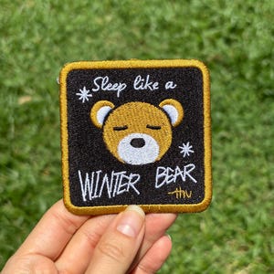 Może przedstawiać: Haftowana naszywka z czarnym tłem i złotą obwódką. Naszywka przedstawia kreskówkowego misia z zamkniętymi oczami i napisem "Sleep like a WINTER BEAR". Dwa białe płatki śniegu są obecne.