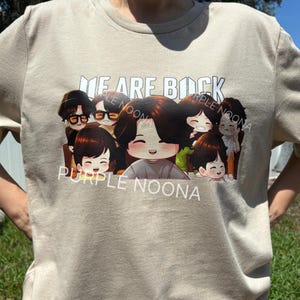 Może przedstawiać: Beżowa koszulka z kreskówkową ilustracją siedmiu uśmiechniętych postaci. Tekst "WE ARE BACK" i "PURPLE NOONA" są wydrukowane na biało. Koszulka wykonana jest z miękkiej tkaniny w neutralnym kolorze.