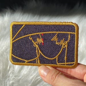 Może przedstawiać: Haftowana naszywka z ciemnofioletowym tłem i złotą obwódką. Naszywka przedstawia złoty rysunek liniowy dwóch postaci z czerwonym akcentem. Naszywka ma kształt prostokąta.