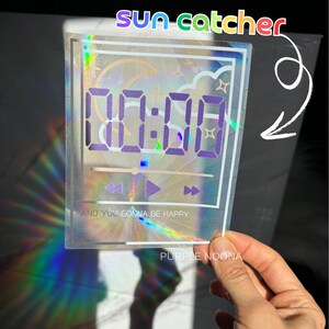 May include: A holographic suncatcher with a digital clock face displaying 00:00, a crescent moon, clouds, and the text "And you gonna be happy". The suncatcher is made of clear acrylic and has a rainbow iridescent finish. The text "Purple Noona" is printed in the bottom right corner.