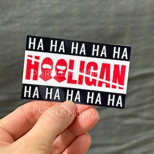Puede incluir: Una pegatina rectangular con la palabra "HOOLIGAN" en rojo y la frase "HA HA HA HA" en blanco sobre un fondo negro. La pegatina también presenta dos gráficos de pasamontañas rojos.
