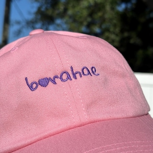 Puede incluir: Una gorra de béisbol rosa con un corazón bordado en morado y la palabra "borahae" en la parte delantera.