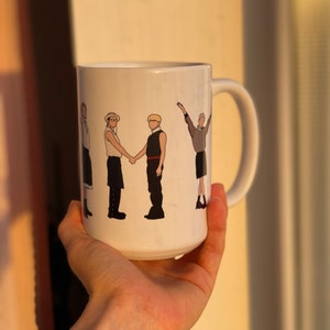 May include: White ceramic mug with a black and white illustration of four people standing in a line. The people are wearing different outfits and have different hairstyles. The mug has a black handle.