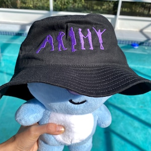 Puede incluir: Sombrero de pescador negro con la palabra "ARMY" bordada en morado. El sombrero lo lleva un peluche azul. El sombrero tiene un ala ancha y está hecho de una tela suave. El peluche es azul claro con una barriga blanca.