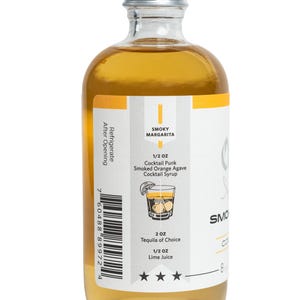 Puede incluir: Una botella de vidrio transparente con tapa plateada, llena de un l&iacute;quido dorado. La etiqueta presenta el texto "SMOKY MARGARITA" y una ilustraci&oacute;n de c&oacute;ctel. La etiqueta tambi&eacute;n enumera ingredientes y medidas, incluyendo "1/2 OZ Cocktail Punk Smoked Orange Agave Cocktail Syrup".