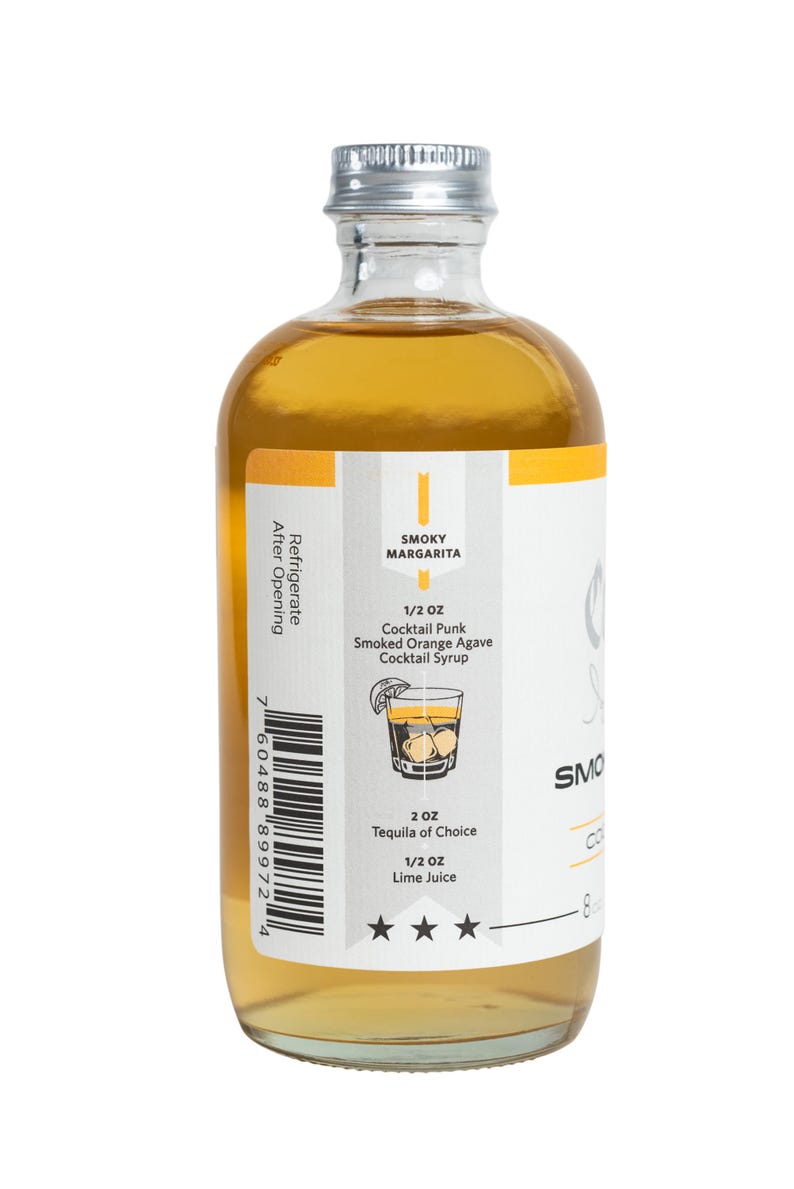 Puede incluir: Una botella de vidrio transparente con tapa plateada, llena de un l&iacute;quido dorado. La etiqueta presenta el texto "SMOKY MARGARITA" y una ilustraci&oacute;n de c&oacute;ctel. La etiqueta tambi&eacute;n enumera ingredientes y medidas, incluyendo "1/2 OZ Cocktail Punk Smoked Orange Agave Cocktail Syrup".