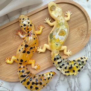 Könnte beinhalten: Zwei Gecko-Figuren aus Harz mit schwarzen und gelben Flecken. Ein Gecko hat einen Schwanz, der zu einem Kreis geformt ist, während der andere einen Schwanz mit einem floralen Design hat.