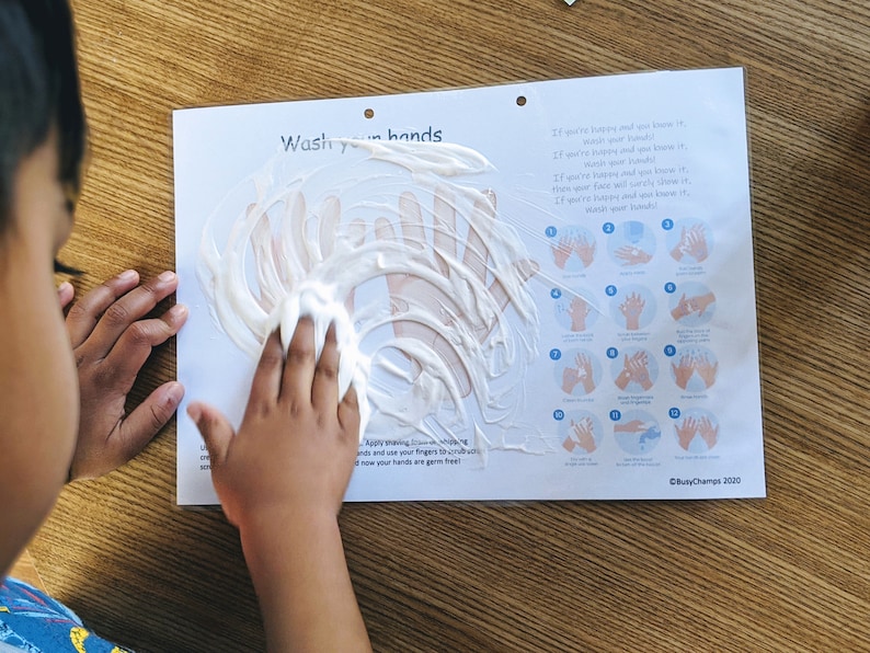 May include: A child's hand is shown applying white foam to a poster with instructions on how to wash hands. The poster has the text "Wash your hands" and a series of illustrations showing the steps for proper handwashing. The poster also includes the text "If you're happy and you know it, wash your hands!" and "Busy Champs 2020."