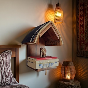 May include: A unique wooden bookshelf shaped like a house, mounted on a wall. The top shelf is formed by an open book, with additional books stacked below. A small decorative elephant figurine sits on the middle shelf. A woven lamp hangs above.