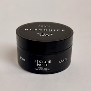 May include: A black jar of hair styling paste labeled "Agate Blacknife Studio Texture Paste Water Base Net: 4 fl.oz/120ml"