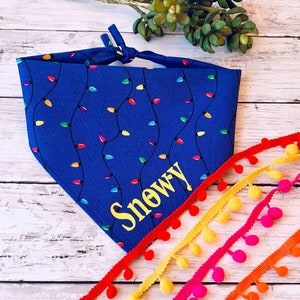 May include: A blue dog bandana with a repeating pattern of colorful Christmas lights. The name "Snowy" is embroidered in yellow. Colorful pom-pom trim in red, yellow, pink, and orange accents the image.