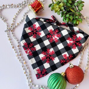 May include: A black and white checkered bandana with red snowflake embroidery. The bandana is surrounded by silver beads, green and red Christmas ornaments, and a small red drum ornament. A sprig of green leaves is also present.
