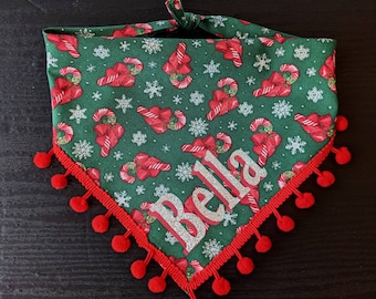 May include: Green Christmas pet bandana featuring red candy canes, bows, and snowflakes. The bandana has a red trim with red pom-poms and the name "Bella" in silver glitter letters.