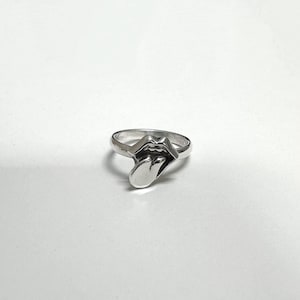 Könnte beinhalten: Ein silberner Ring mit einem stilisierten offenen Mund mit herausgestreckter Zunge. Der Ring besteht aus glänzendem Metall und ist vor einem schlichten weißen Hintergrund platziert. Das Design ist ein auffälliges Schmuckstück.