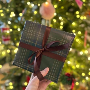Peut inclure: Un cadeau emball&eacute; dans du papier &agrave; carreaux vert et un ruban marron nou&eacute; en n&oelig;ud. L'arri&egrave;re-plan est un sapin de No&euml;l flou avec des lumi&egrave;res et des d&eacute;corations.