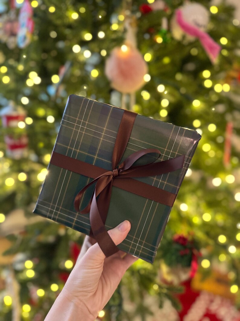 Peut inclure: Un cadeau emball&eacute; dans du papier &agrave; carreaux vert et un ruban marron nou&eacute; en n&oelig;ud. L'arri&egrave;re-plan est un sapin de No&euml;l flou avec des lumi&egrave;res et des d&eacute;corations.