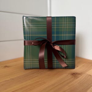 Peut inclure: Un cadeau emball&eacute; dans du papier vert &agrave; carreaux, orn&eacute; d'un ruban de satin marron fonc&eacute; nou&eacute; en un n&oelig;ud. Le cadeau est pos&eacute; sur une surface en bois.