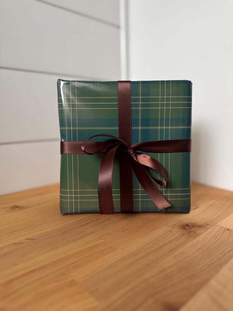 Peut inclure: Un cadeau emball&eacute; dans du papier vert &agrave; carreaux, orn&eacute; d'un ruban de satin marron fonc&eacute; nou&eacute; en un n&oelig;ud. Le cadeau est pos&eacute; sur une surface en bois.