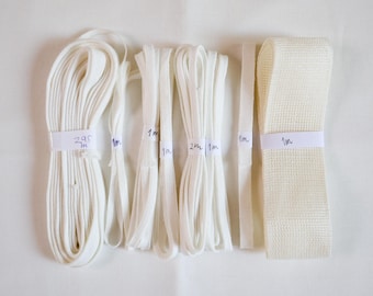 May include: A collection of white elastic bands and fabric tapes arranged in various lengths. The items are bundled with paper labels marked with measurements including 295m, 1m, 2m, and 1m. The assortment includes thin cords and a wide, textured webbing strap.