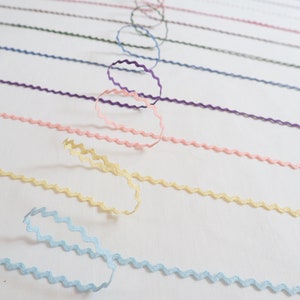 Może przedstawiać: Zestaw kolorowych wstążek z falbanką w różnych kolorach, w tym jasnoniebieski, żółty, różowy, fioletowy i zielony. Wstążki ułożone są w rzędach na białym tle.