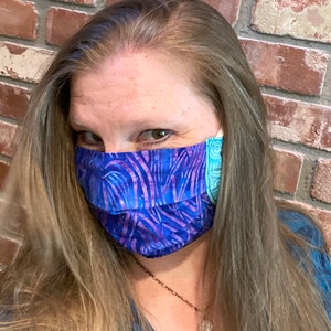 May include: A woman wearing a blue and purple patterned face mask with a turquoise and white patterned border. The mask is covering her nose and mouth. She has long brown hair and is wearing a blue shirt.