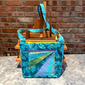 May include: A turquoise and blue fabric crossbody bag with a palm leaf pattern. The bag features a geometric design with shades of blue and green, a yellow zipper, and a purple zipper pull. The adjustable strap is also turquoise and blue.
