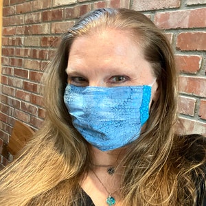 May include: A woman wearing a blue patterned face mask with a white background. The mask has a blue and white polka dot pattern on the bottom edge. She is wearing a black shirt and a silver necklace with a turquoise pendant.