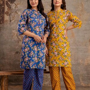 May include: Two matching sets consisting of a long-sleeved tunic and trousers. One set is blue with a pink and white floral pattern, and the other is yellow with a similar floral print. Both tunics feature a mandarin collar and a buttoned placket.