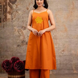 May include: A sleeveless orange midi dress with thin straps and a yellow star-shaped embroidered patch on the chest, paired with matching wide-leg orange trousers. The outfit features a solid colour design with a simple neckline and a relaxed fit.